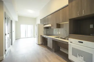 The inside of a vacant apartment unit. There is a kitchen and empty living space with patio doors. Bright natural light is coming in the window.