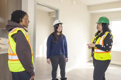 Three people stand in a group inside an empty Waterloo Region Housing unit. Two people are wearing safety vests. Two people are wearing hardhats for construction.