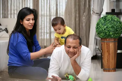 An older baby crawling on their dad's back while he is reading, with the mother holding the baby's arm as they crawl.
